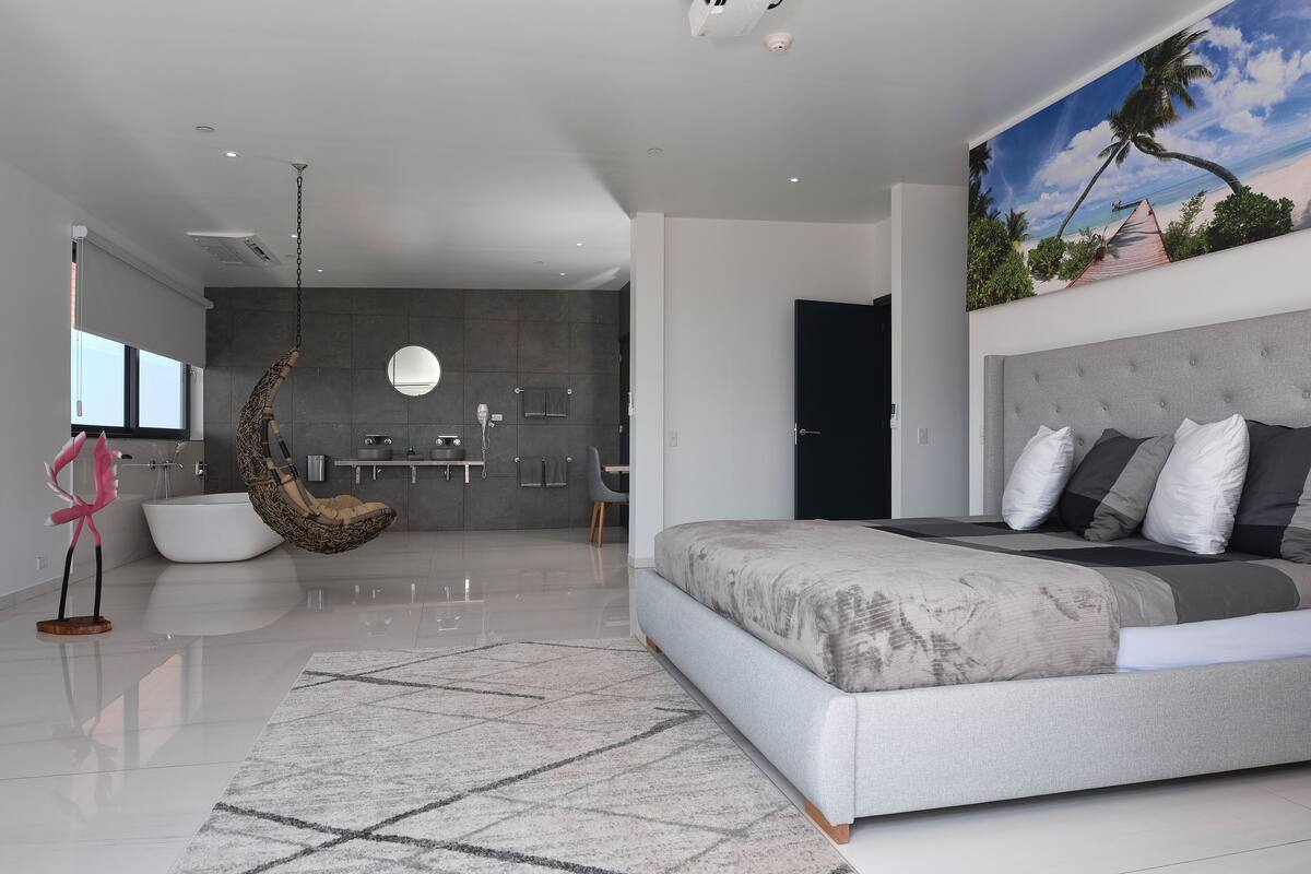 A modern bedroom with a large bed, a hanging chair, and a view of a bathroom and kitchen.