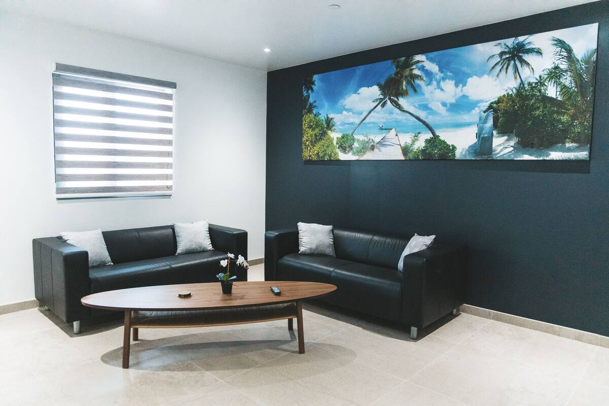 A modern living room with two black leather sofas, a wooden coffee table, and a large tropical-themed painting on the wall.