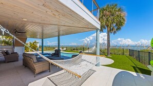 A covered outdoor area with a hammock, wicker furniture, and a view of a pool and palm tree. - Sea La Vie AM: Bay front, pool, hot tub, rooftop deck, and water sports gear! (Anna Maria)
