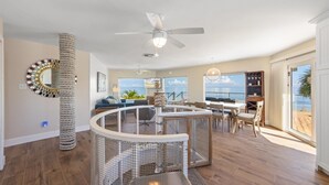 A spacious living area with a high ceiling fan, a circular play area for pets, a dining table with chairs, and a view of the ocean through large windows. - Sea La Vie AM: Bay front, pool, hot tub, rooftop deck, and water sports gear! (Anna Maria)