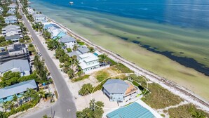 An aerial view of a coastal residential area with houses along the beachfront, a clear blue sea, and a boat in the distance. - Sea La Vie AM: Bay front, pool, hot tub, rooftop deck, and water sports gear! (Anna Maria)