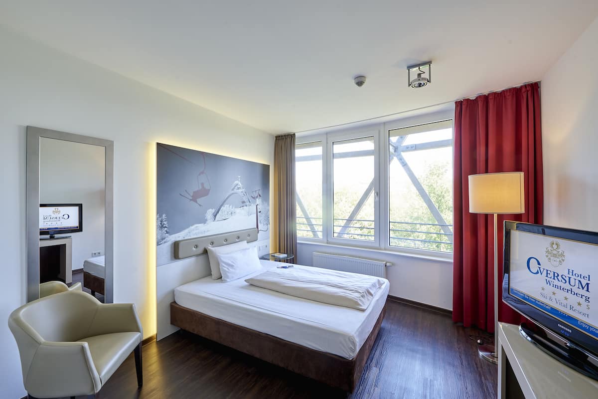 A hotel room with a bed, a TV, a desk with a computer, a chair, and a window with curtains.