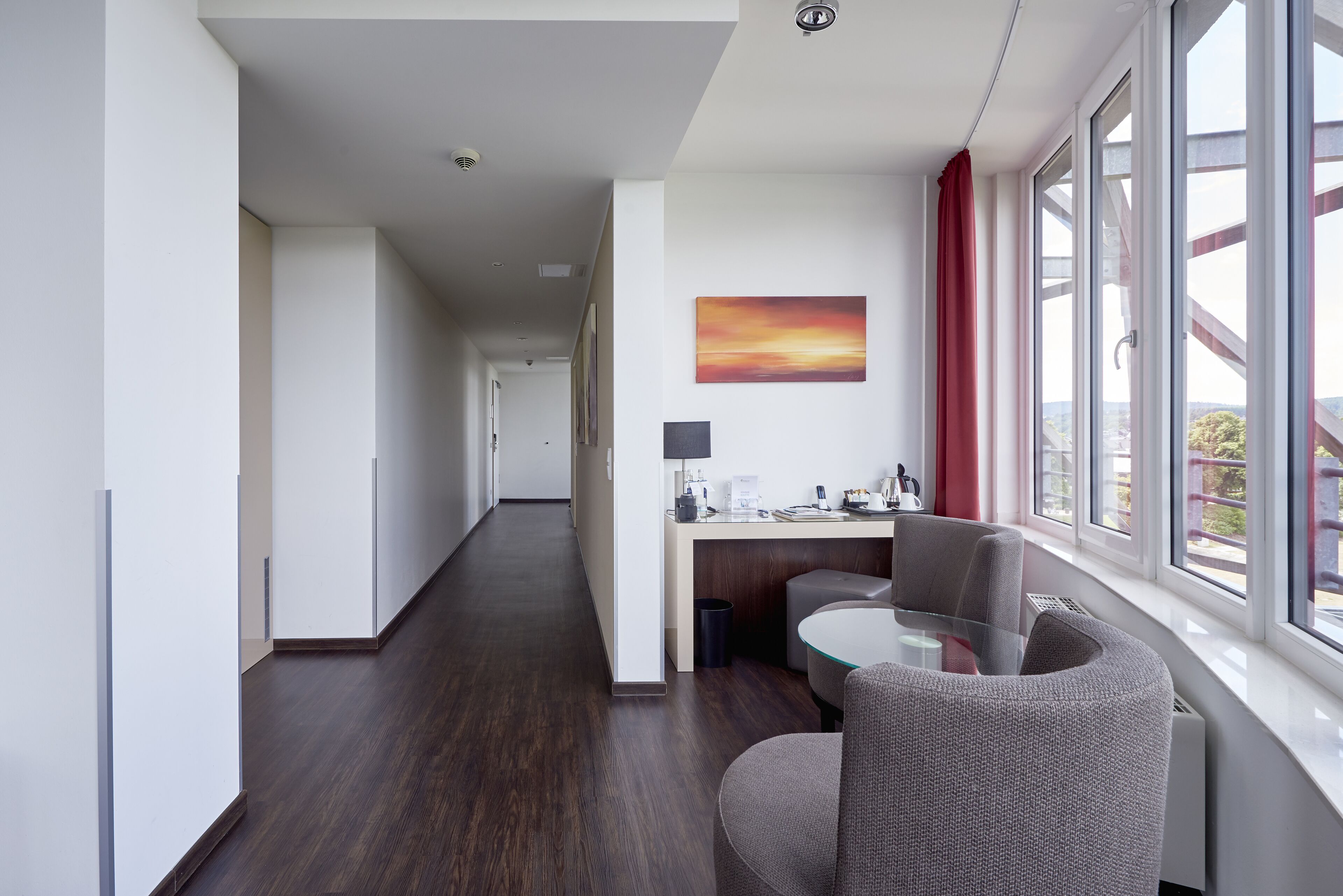 Habitació d'hotel moderna amb un llarg passadís, zona de menjador amb taula de vidre i una finestra gran amb cortina vermella.