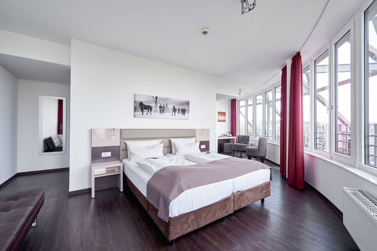 A modern hotel room with a large bed, a desk, and a chair. There is a window with curtains and a view of a bridge.