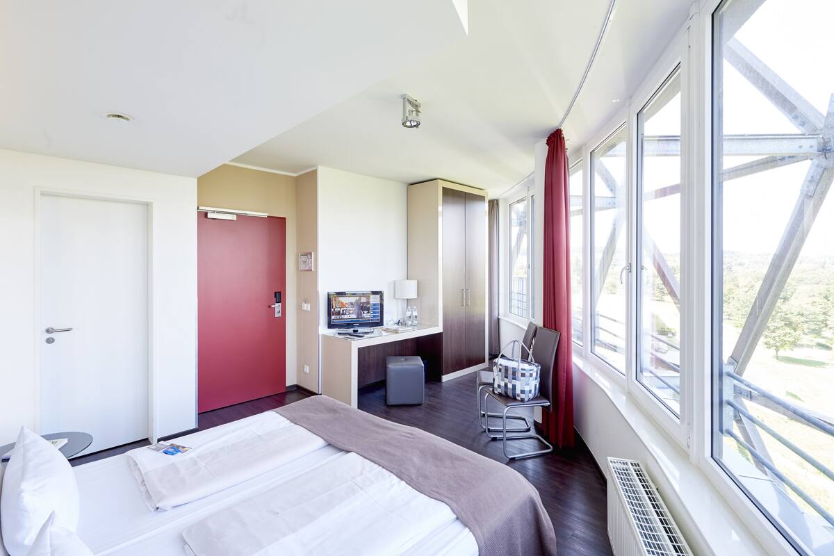 A hotel room with a bed, desk, chair, and large windows.