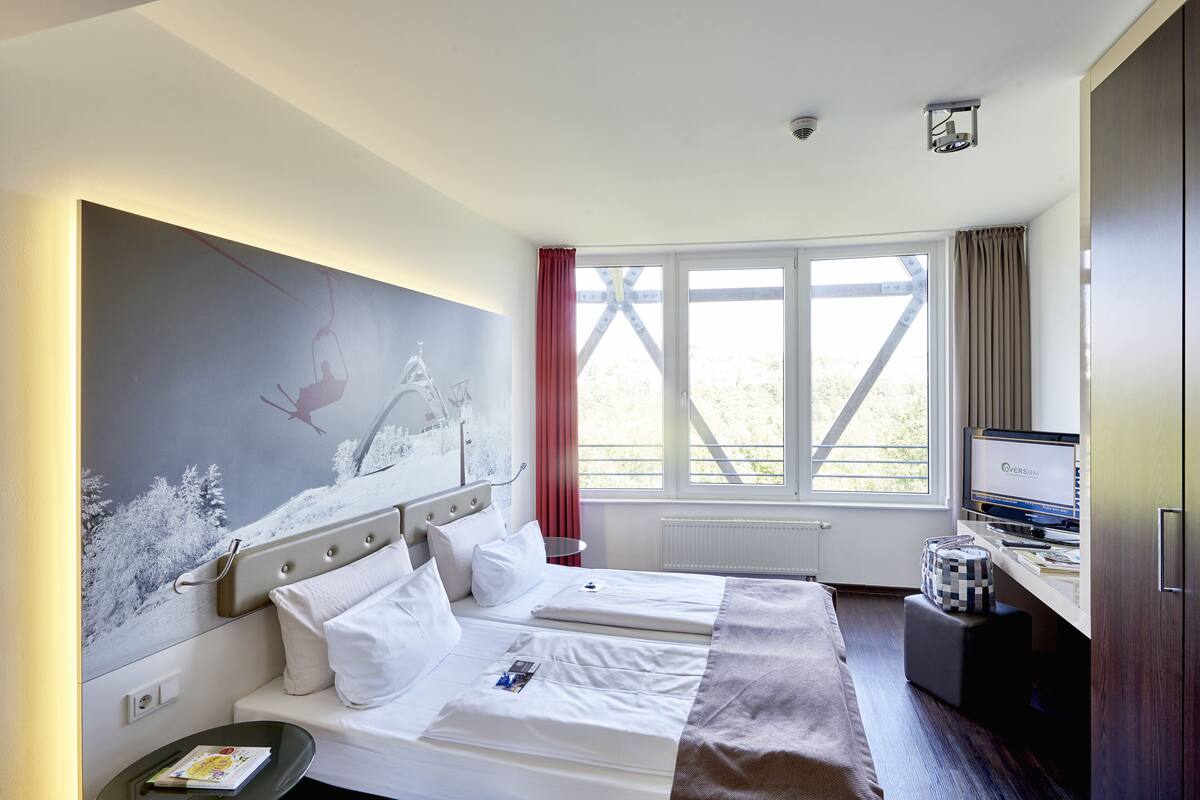 A hotel room with two beds, a large window with curtains, a desk with a computer, and a wall-mounted artwork.