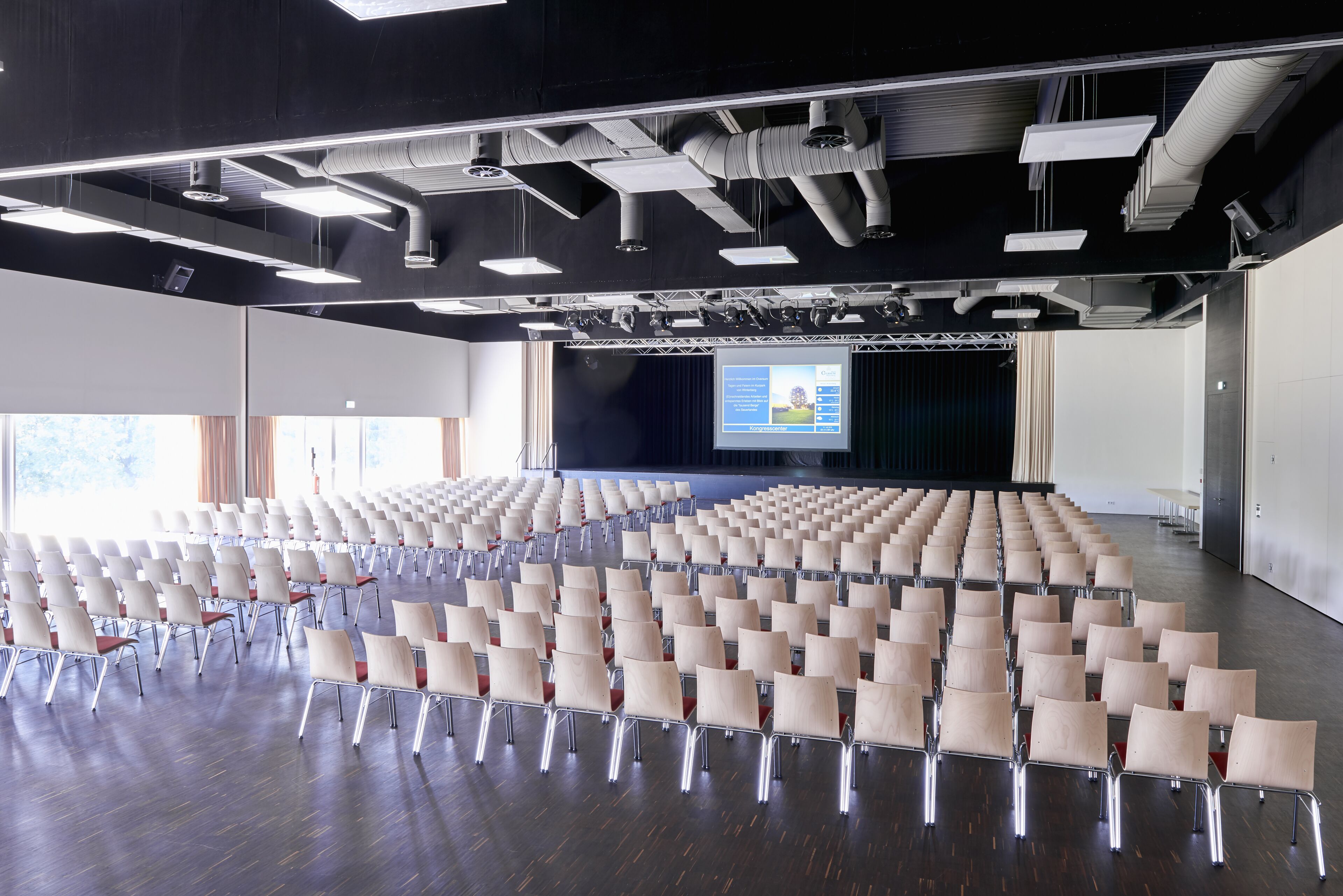 Un salón de conferencias con filas de sillas, un escenario con una pantalla de presentación y un amplio ventanal que proporciona luz natural.