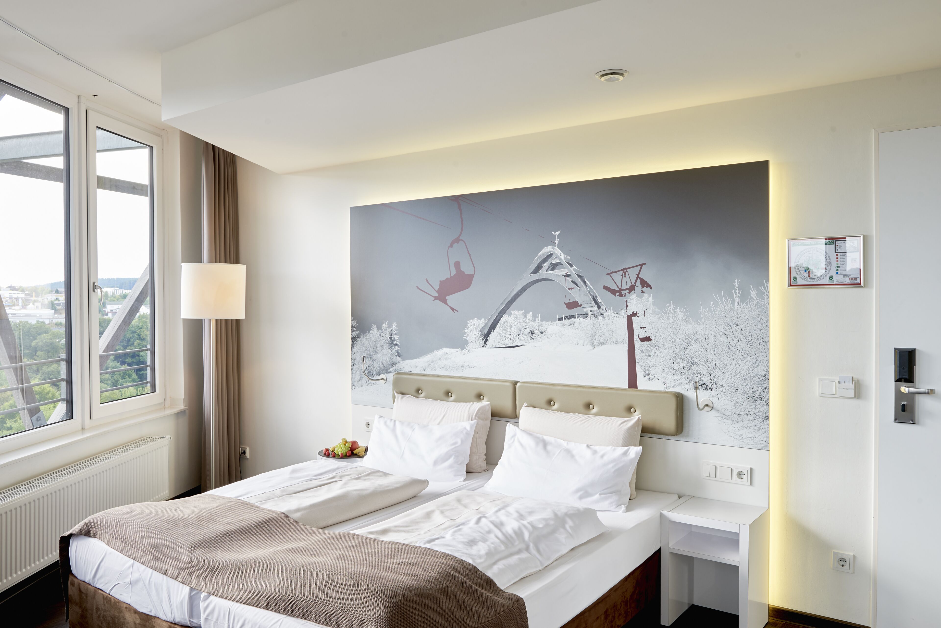 A hotel room with a large bed, a view of the city through the window, a painting on the wall, and a nightstand with a lamp.
