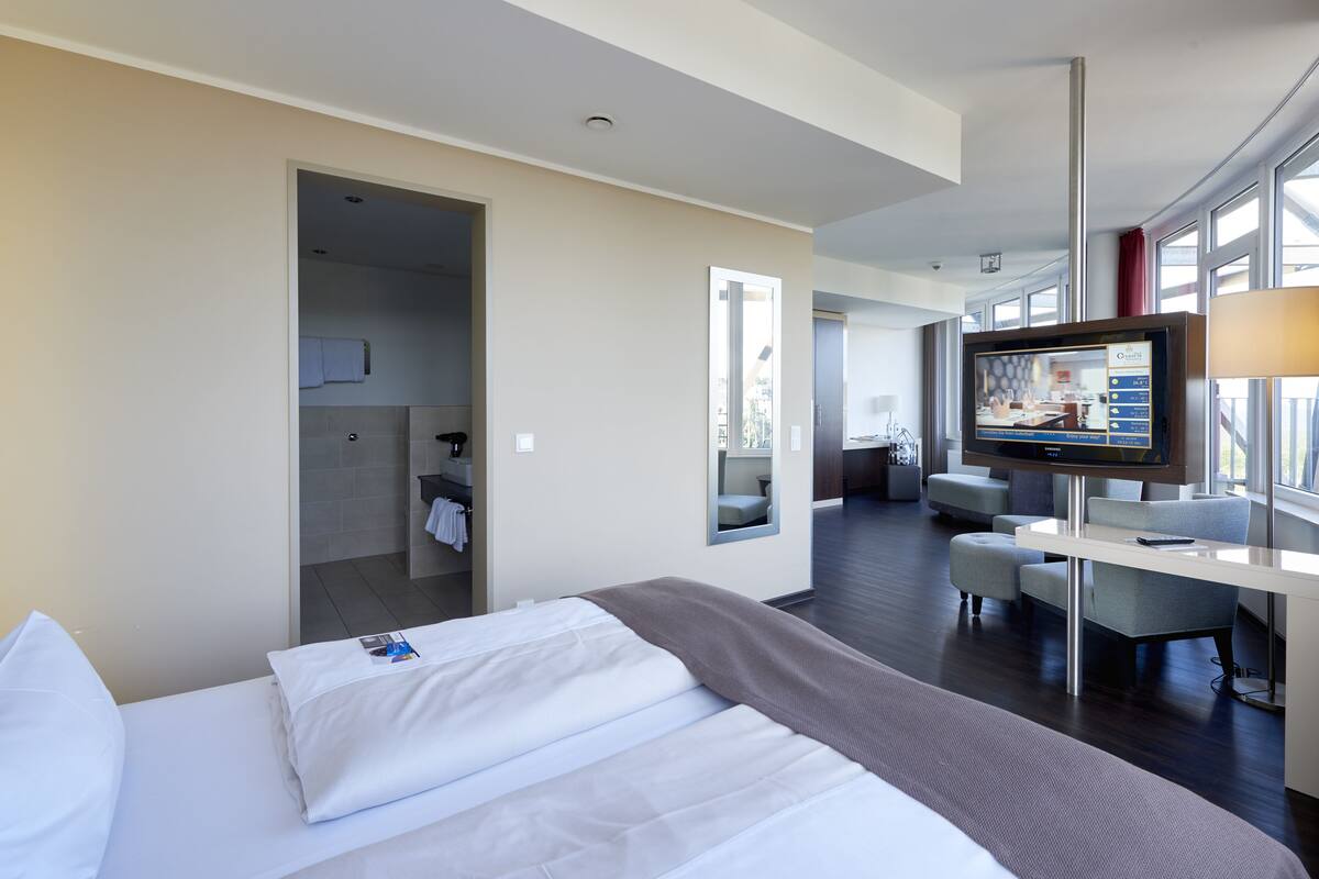 A modern hotel room with a large bed, a desk with a flat-screen TV, and a view of the city through large windows.