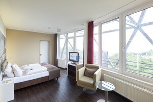 Ein Hotelzimmer mit einem Bett, einem Schreibtisch mit Computer, einem Stuhl und großen Fenstern mit Blick auf eine Brücke.