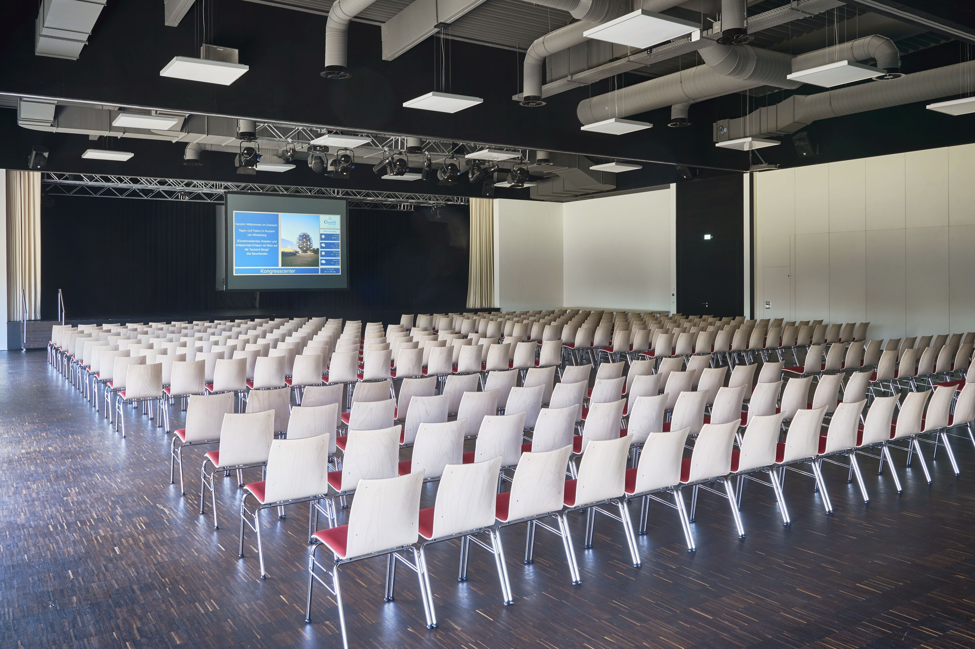 Un amplio salón de conferencias con filas de sillas blancas y una pantalla de proyección.