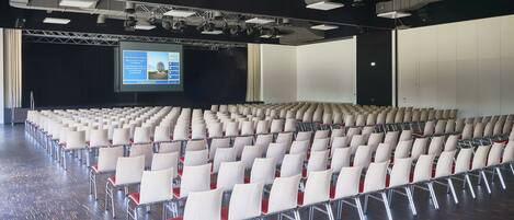Un amplio salón de conferencias con filas de sillas blancas y una pantalla de proyección.
