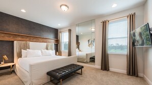 A modern hotel bedroom with a large bed, a bench, a bedside table, a mirror, and a window with curtains.