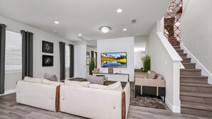 A modern living room with a large flat-screen TV, a sofa, and a staircase. - Striking Home near Disney: Games, Outdoor Kitchen & Waterpark - 7742FF (Kissimmee)