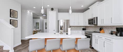 Una cocina moderna con armarios blancos, una isla central con taburetes y electrodomésticos de acero inoxidable, además de iluminación colgante.