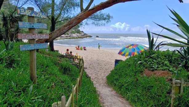 Un sendero bordeando la playa que lleva al océano, con una sombrilla de colores y gente a lo lejos.