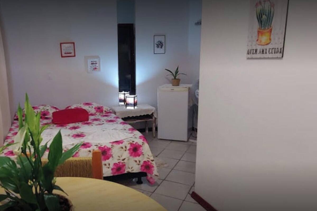 Quarto com cama, escrivaninha, cadeira, planta em vaso e um pequeno frigorífico.
