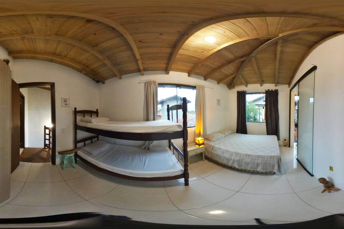 Quarto com cama de solteiro, teto de madeira, janela com cortinas e guarda-roupa.