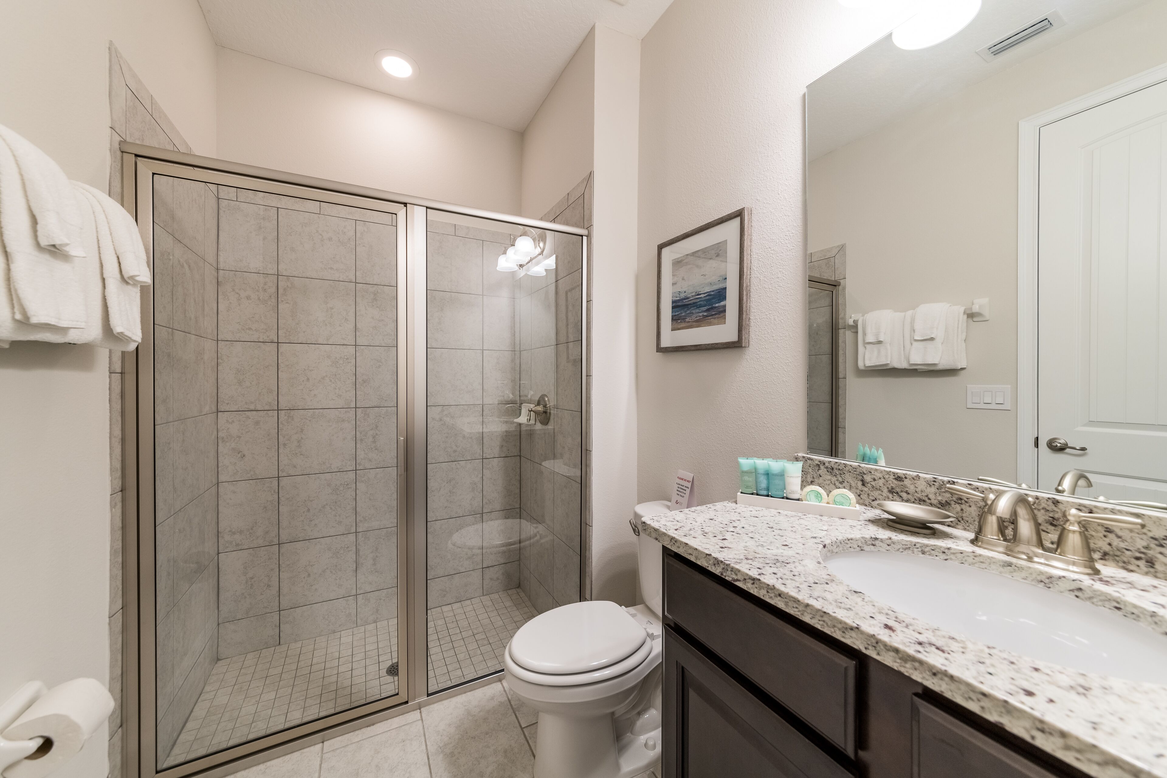 A bathroom with a glass-enclosed shower, a white toilet, a sink with a granite countertop, and a mirror above the sink.