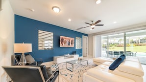 A modern living room with a blue accent wall, a ceiling fan, a glass coffee table, a white sofa, and a large window offering a view of the outdoors. - Magnificent Home near Disney: Theater, Pool Table & Waterpark - 7448MR (Reunion)