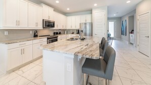 A modern kitchen with white cabinets, a marble countertop, stainless steel appliances, and a bar area with seating. - Magnificent Home near Disney: Theater, Pool Table & Waterpark - 7448MR (Reunion)
