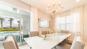 A dining area with a glass table, cushioned chairs, a chandelier, and a view of a swimming pool. - Luxury Home near Disney: Themed Rooms, Air Hockey & Waterpark - 7699GR (Reunion)