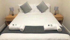 Un lit bien fait, avec du linge de lit blanc, deux oreillers gris et une tête de lit grise. On y trouve deux tables de chevet avec des lampes, ainsi qu’une petite table de nuit en bois, également avec une lampe.