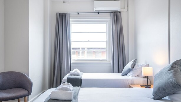 Une chambre d’hôtel avec deux lits, une chaise, une fenêtre avec des rideaux, une lampe et un appareil de climatisation.