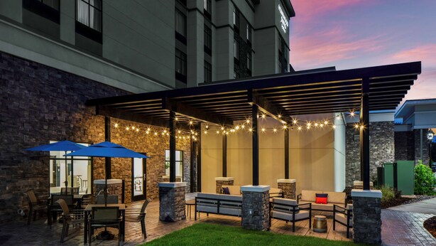 An outdoor patio area with stone pillars, blue umbrellas, and string lights. - Homewood Suites by Hilton Greensboro Wendover, NC (Greensboro)