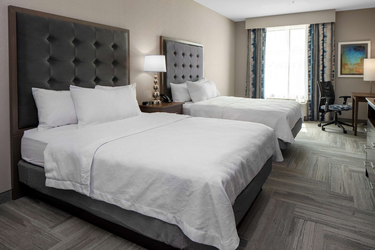 A hotel room with two beds, a desk, a chair, and a large window with curtains.