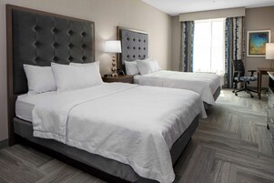 Habitación de hotel con dos camas, un escritorio, una silla y un amplio ventanal con cortinas.