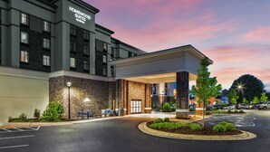 A hotel entrance with a stone facade, a sign reading "Homewood Suites by Hilton," and a parking lot with cars and a landscaped area. - Homewood Suites by Hilton Greensboro Wendover, NC (Greensboro)