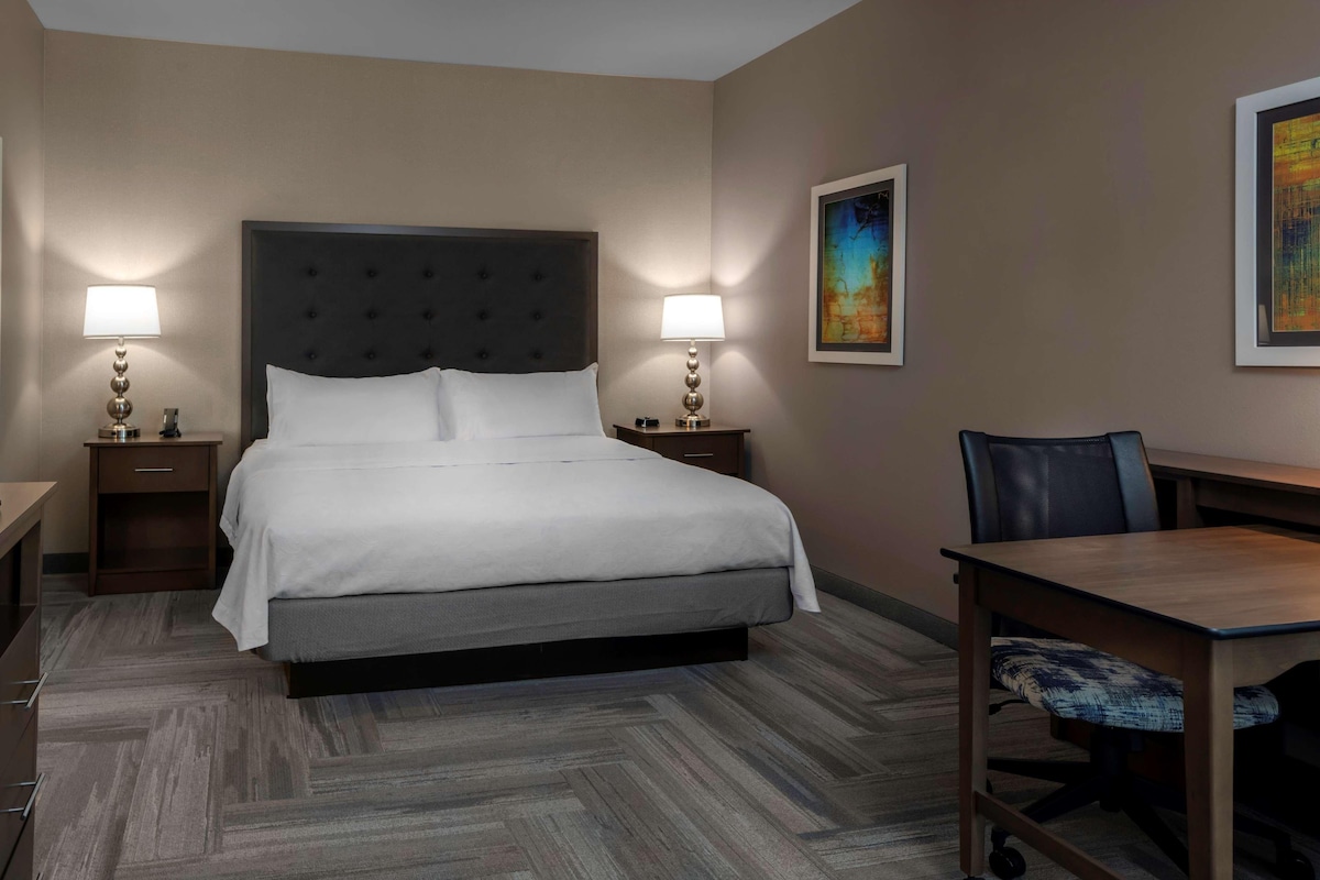Habitación de hotel con una cama grande, dos mesitas de noche con lámparas, un escritorio con una silla y dos cuadros enmarcados en las paredes.