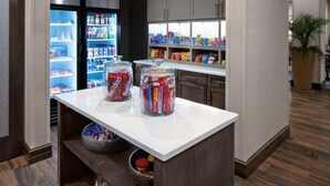A kitchen area with a refrigerator stocked with beverages, a countertop with jars of snacks, and cabinets above. - Homewood Suites by Hilton Greensboro Wendover, NC (Greensboro)