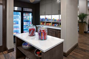 Una zona de cocina con un refrigerador lleno de bebidas, una encimera con frascos de aperitivos y alacenas arriba.