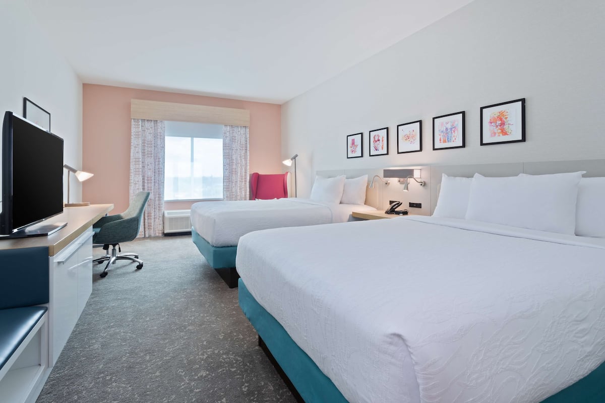 A hotel room with two beds, a desk with a TV, a chair, and a window with curtains.