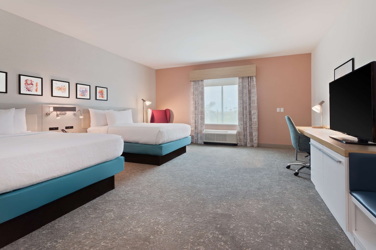 A hotel room with two beds, a desk with a TV, and a window with curtains.