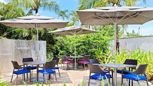 Un espace extérieur aménagé en terrasse, avec plusieurs tables et chaises, entouré d’une végétation luxuriante et de palmiers.