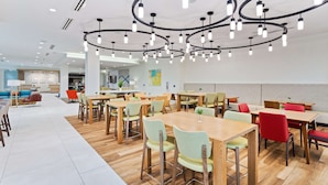 Un espace de restauration moderne, avec des tables et des chaises en bois, un lustre central et une ambiance lumineuse et aérée.