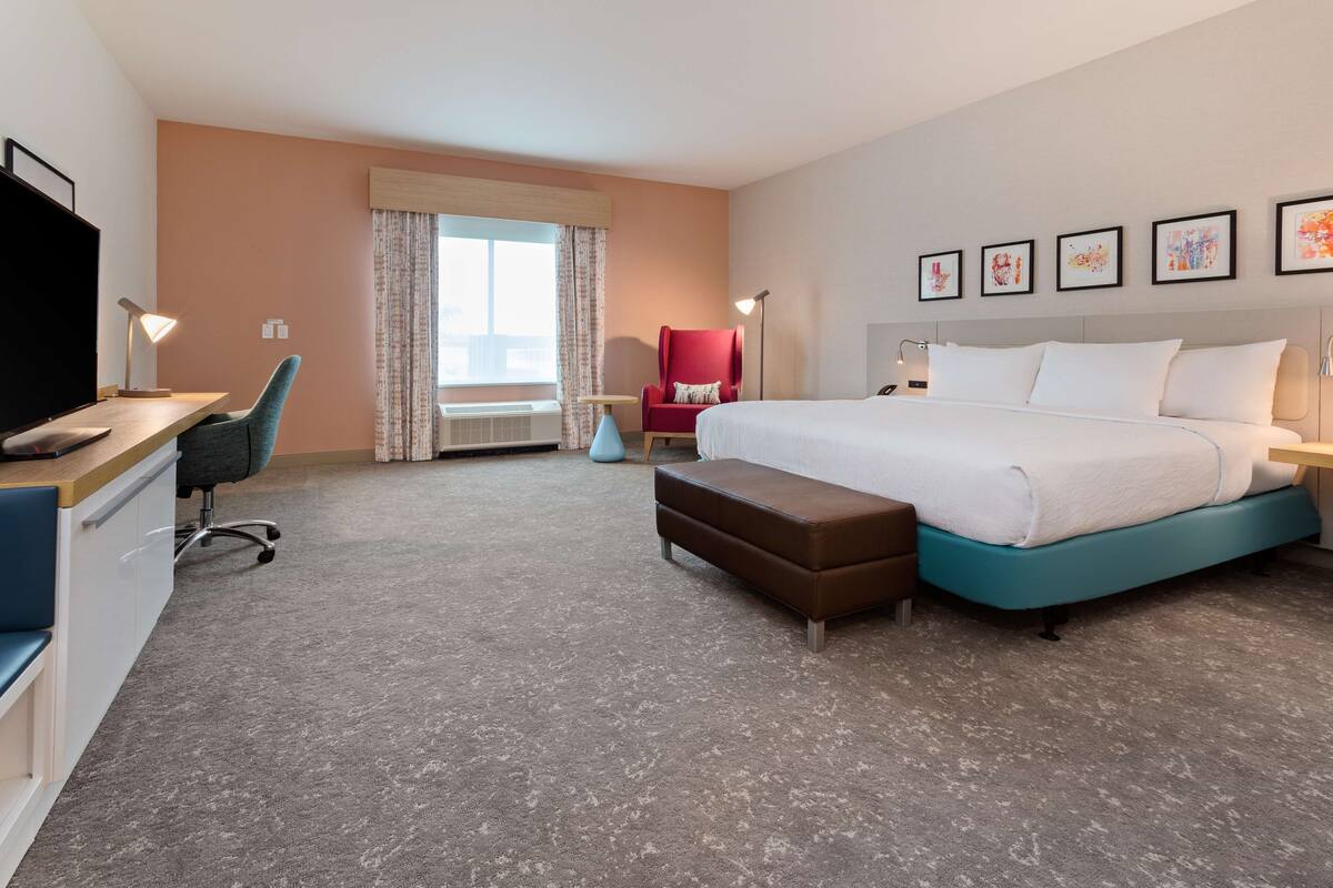 A hotel room with a bed, desk, chair, and artwork on the wall.