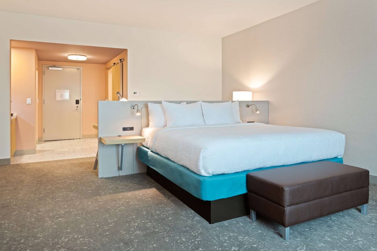 A modern hotel room with a bed, bedside tables, a bench, and a door leading to a hallway.