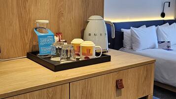 A hotel room with a wooden dresser, a white coffee maker, a tray with mugs and a bottle, and a bed with white pillows.