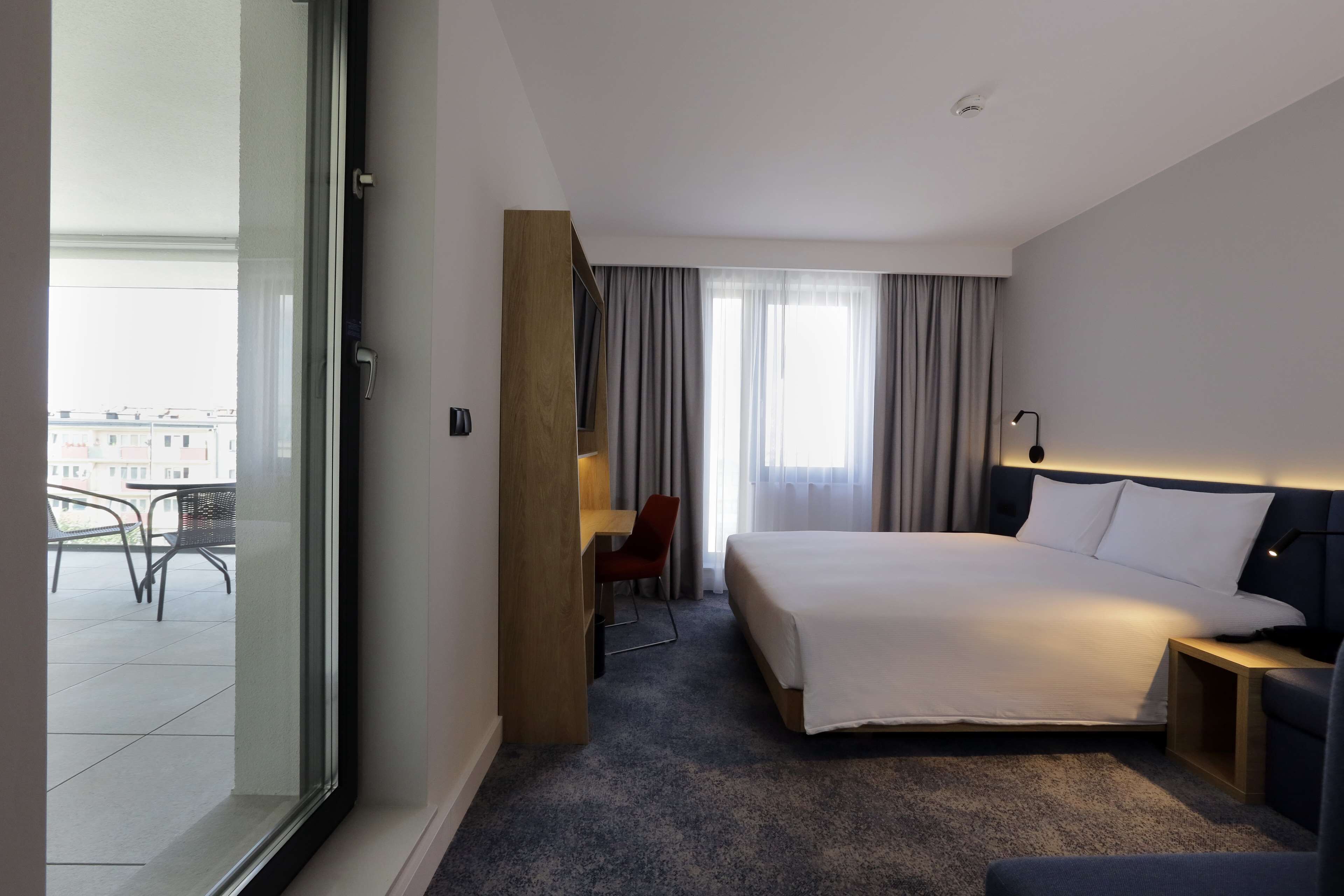 A hotel room with a large bed, a desk, a chair, and a view of the city through the window.