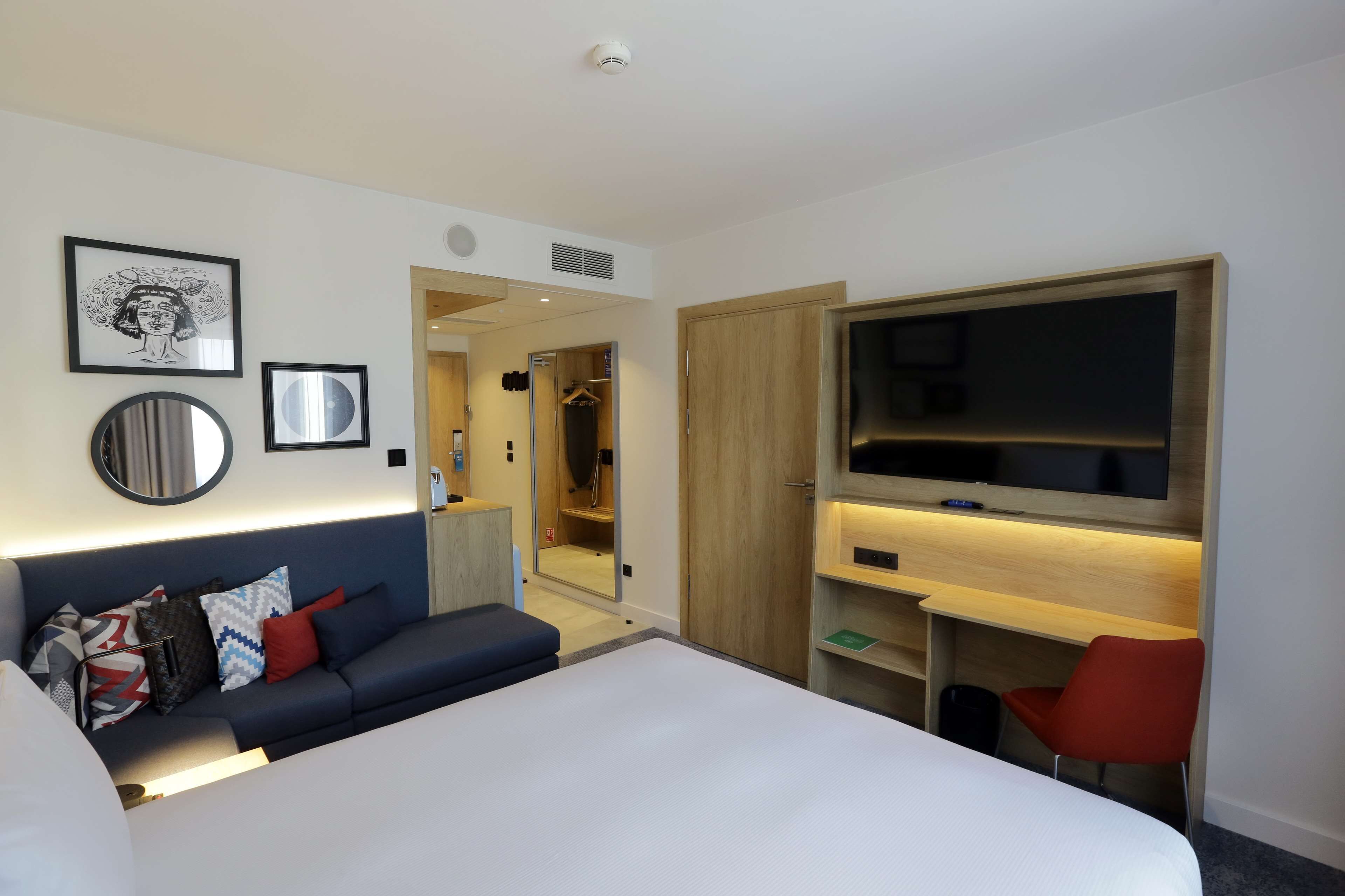 A modern hotel room with a bed, a desk, a TV, a mirror, and a wardrobe.