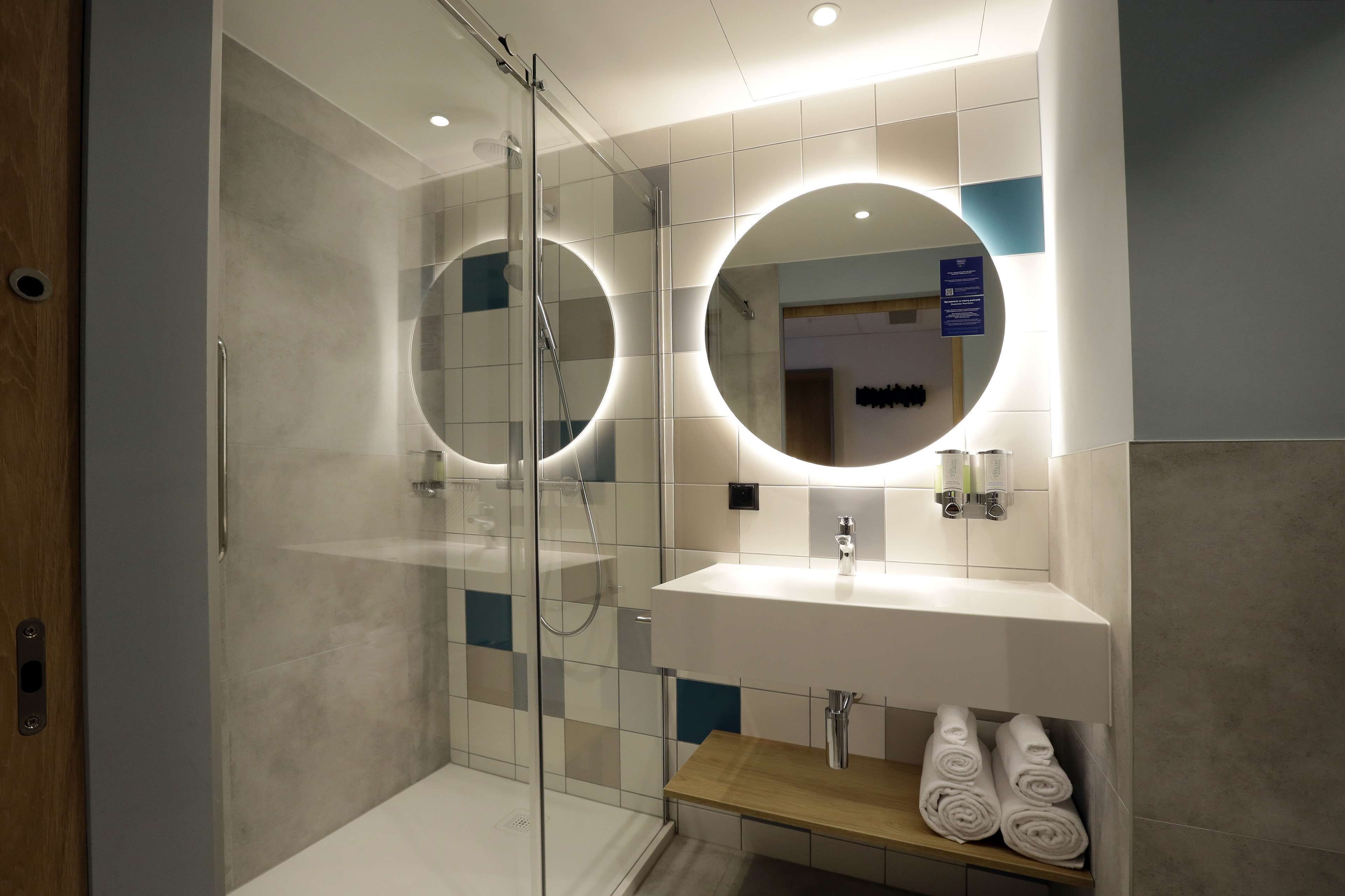 Un baño moderno con ducha envuelta en vidrio, un lavamanos blanco con espejo y un estante con toallas.
