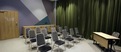 Un salón de conferencias con una larga mesa, filas de sillas, un atril y un telón verde de fondo.
