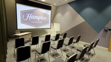 A conference room with a large screen displaying "Hampton by Hilton," arranged chairs, a whiteboard, and a table.