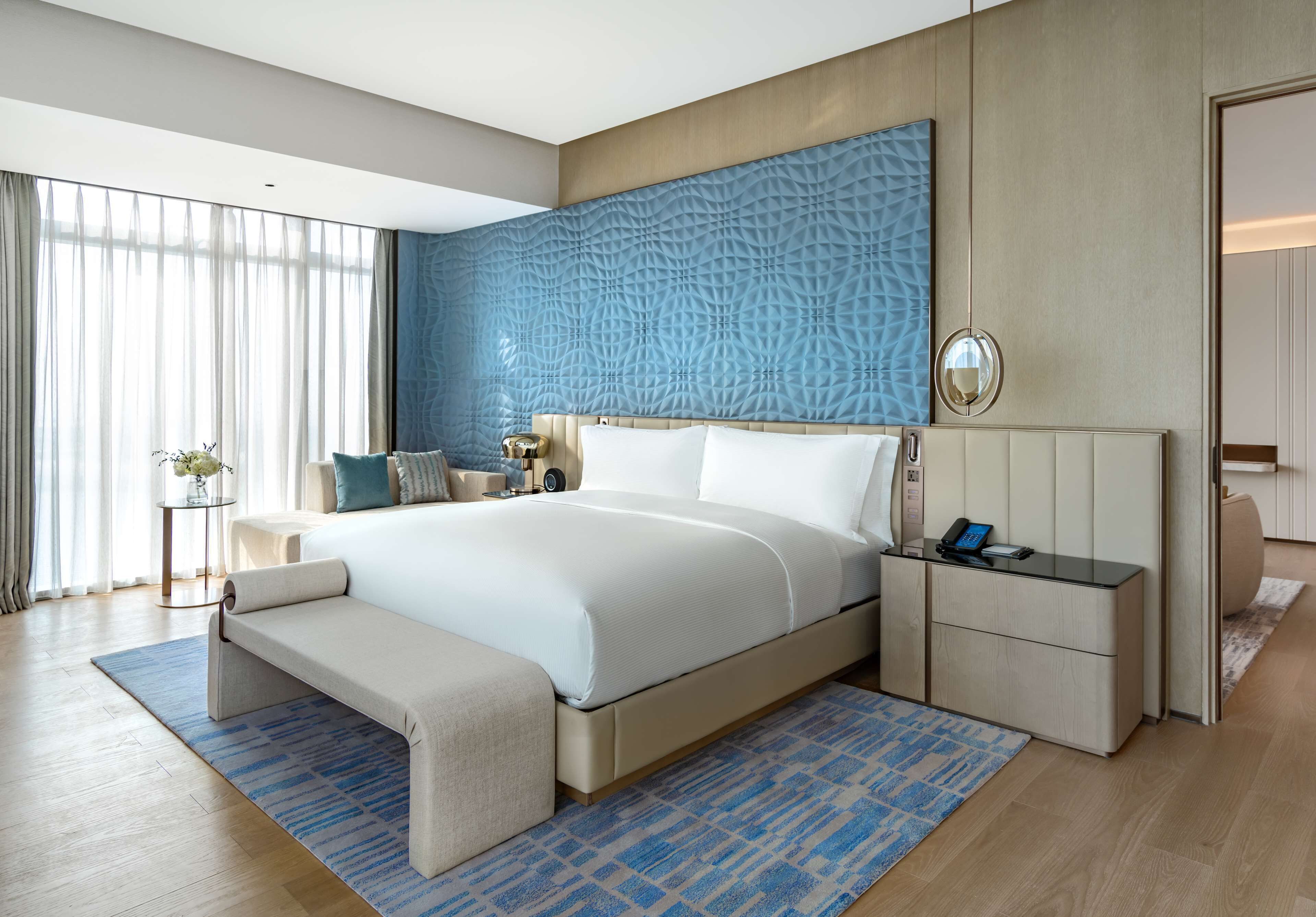 A modern hotel room with a large bed, a bedside table, a nightstand, and a large window with sheer curtains.