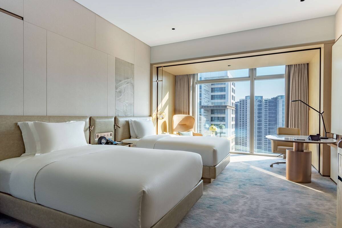 A modern hotel room with a large bed, a desk, and a view of tall buildings.