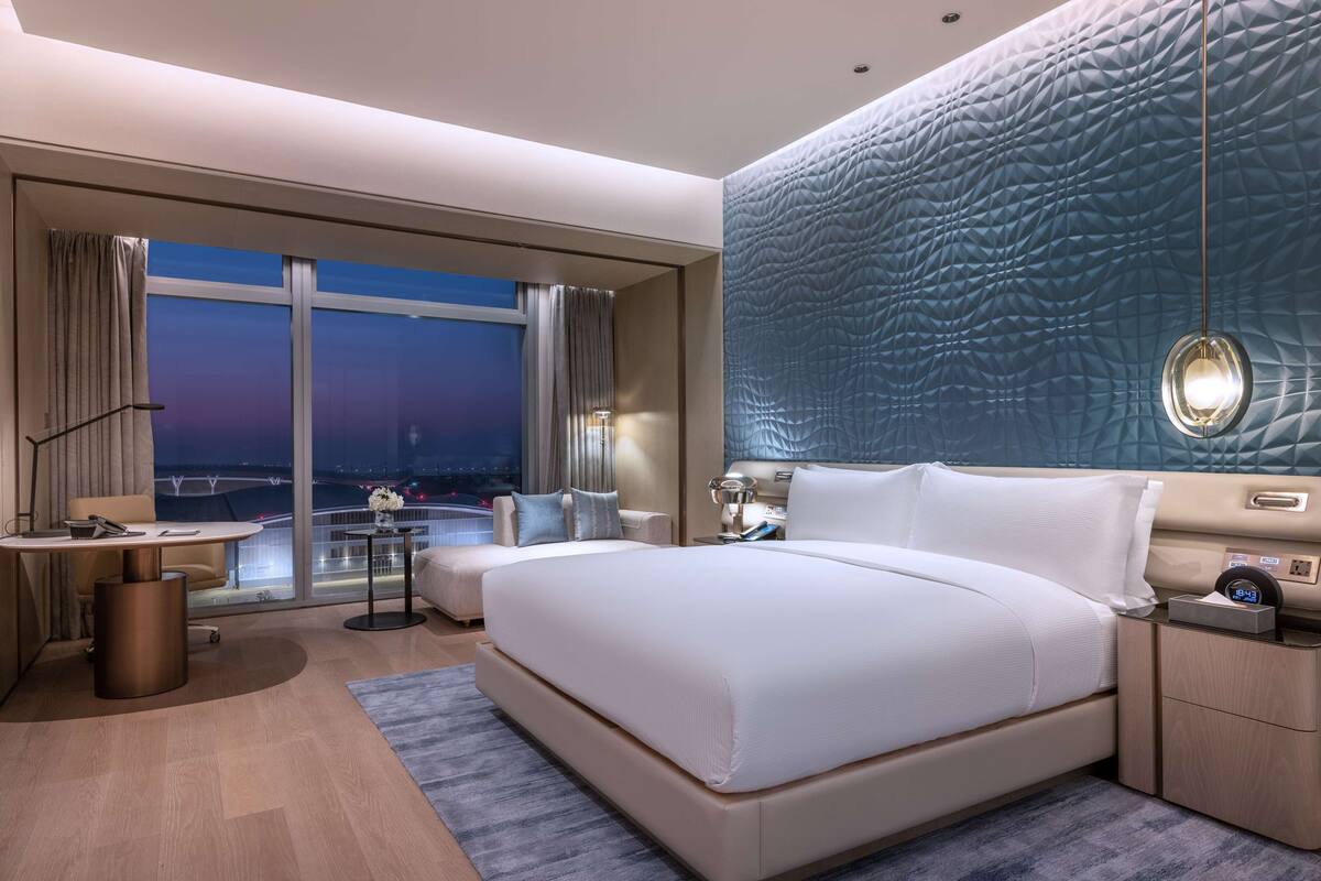 A modern hotel room with a large bed, a desk, a chair, and a view of the cityscape through large windows.